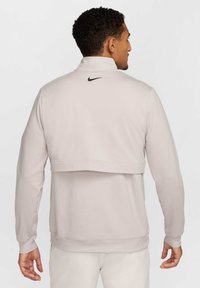 Svetlo šedý mikina Nike s vysokým golierom, prekrývajúcou zadnou časťou, rebrovanými manžetami a malým čiernym logom na krku. Hladká textúra, voľný strih.