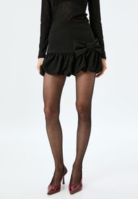 Schwarze Shorts mit gerafftem Saum und großer Schleifendetail an der Taille. Das Modell trägt durchsichtige schwarze Strumpfhosen und glänzende burgunderfarbene Absätze.