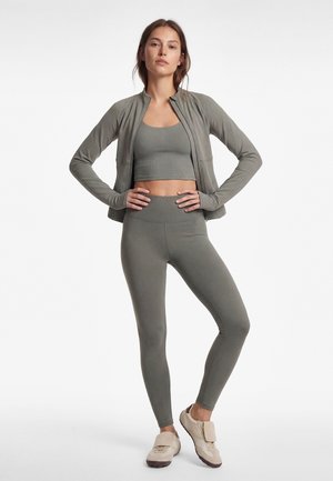 Frau steht mit den Händen in der Hüfte, trägt eine olivgrüne Jacke mit Reißverschluss, ein passendes Croptop und Leggings sowie beige Turnschuhe vor weißem Hintergrund.