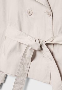 Veste en tissu beige clair avec une ceinture nouée à la taille et deux boutons visibles près du col.