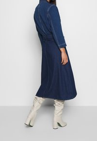 Vestido camisero de mezclilla en azul oscuro, de manga larga, con cintura ceñida y falda completa. Presenta costuras contrastantes y una silueta fluida. Se combina con botas crema hasta la rodilla.
