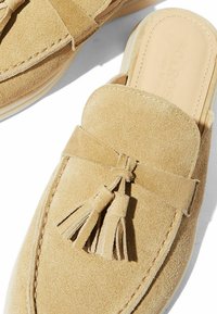 Mocassini in suede beige con punta arrotondata, dotati di una larga fascia, dettaglio con nappina e una suola in gomma chiara. Intersuola in pelle liscia.