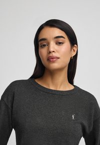 Dunkelgrauer Strickpullover mit Rundhalsausschnitt und gerippten Bündchen, der ein kleines gesticktes Detail auf der linken Brust hat. Weiche Textur.