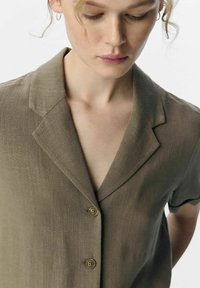 Camisa de manga corta verde oliva con cuello acentuado, textura ligera y dos botones marrones en el frente.