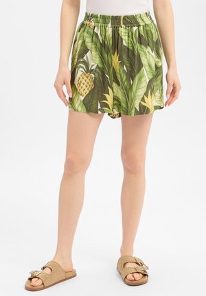 Femme portant un short imprimé feuilles tropicales et ananas et des sandales beige sans lacets, debout devant un fond blanc.
