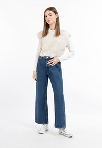 Vesta tricotată bej peste un top cu mâneci lungi, asortată cu blugi albastre cu talie înaltă și picioare largi, purtată cu adidași albi, pe un fundal simplu.