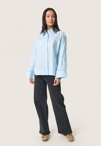 Lichtblauwe longsleeve shirt met een button-down kraag en decoratieve stiksels, gecombineerd met donkere wijde jeans en beige schoenen.