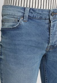 Jeans de mezclilla en un azul claro con un acabado deslavado, diseño de cinco bolsillos y herrajes plateados. Presenta costuras en contraste a lo largo de las costuras.