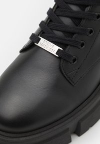 Botte en cuir noir avec semelle en caoutchouc épaisse, lacets noirs passés à travers des œillets, et une étiquette en métal sur les lacets gravée de "STEVE MADDEN".