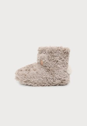 Zapatilla suave, marrón claro, peluda hasta el tobillo con una borla en la parte trasera y pequeñas costuras decorativas que imitan la cara de un oso.