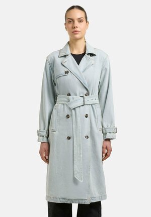 Kvinne iført en lys blå dobbelspent denim trenchcoat med belte, knappede mansjetter og sidelommer over svarte bukser.