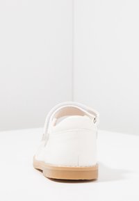 Białe lakierowane buty skórzane z okrągłym czubkiem, rzepem i jasno-brązową gumową podeszwą; widok z tyłu pokazuje wycięty wzór i detale przeszycia.