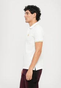 Hvit polo skjorte med gult stripet krage og ermer, med en subtil logo-patch. Korte ermer, sideslisser og myk tekstur.