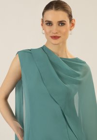 Ein ärmelloses teal-farbenes Kleid aus leichtem Stoff, mit drapierten Designelementen und einer fließenden Silhouette. Subtiler Glanz und weiche Textur.