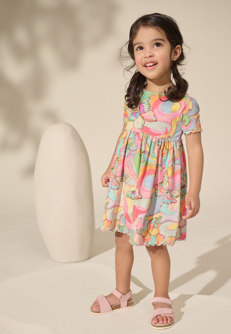 Young girl with dark hair in pigtails wearing a colorful butterfly-patterned dress and pink sandals, standing against a neutral background.