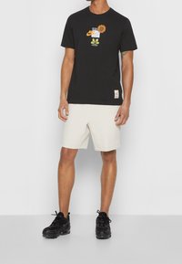 Man i svart Nike-t-shirt med solros och vita shorts, som står med svarta sneakers mot en neutral bakgrund.