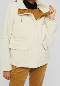 Kermanvärinen fleece-takki, jossa on korkea kaulus, vetoketju, kaksi etutaskua ja vaaleanruskeita yksityiskohtia. Päällä ruskea teksturoitu neule.