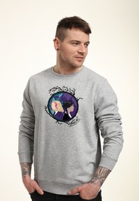 Mu�ž s krátkými vlasy a náušnicemi v uších, oblečený v šedé mikině s uměleckým designem dvou siluet žen obklopených větvemi.