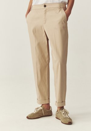 Béžové chinos s manžetami nosené s khaki semišovými teniskami a bielymi šnúrkami, ruky vreckách, na jednoduchom neutrálnom pozadí.