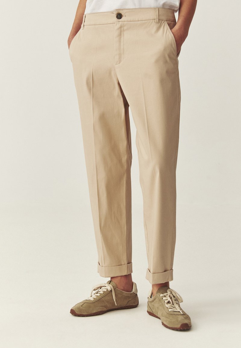 Beige chinos med opbuk ved anklerne, båret med khaki ruskindsneakers med hvide snørebånd, hænderne i lommerne, foran en ensfarvet neutral baggrund.