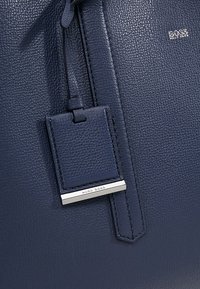 Ciemnoniebieska torebka ze skóry teksturowanej z przymocowaną metką bagażową, z metalową srebrną belką i małym logo "BOSS Hugo Boss" z przodu.