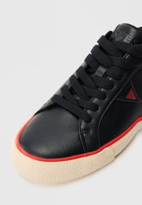 Μαύρο αθλητικό παπούτσι από δέρμα με υφή, με μαύρα κορδόνια, κόκκινες λεπτομέρειες και παχύ, κρεμ χρώματος πάτο. Ορατό το τρίγωνο λογότυπο.