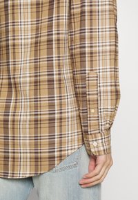 Camicia a maniche lunghe a quadri marrone e beige con polsini a bottoni e bordo curvo, caratterizzata da una morbida trama di cotone e strisce bianche a contrasto.