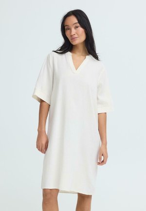 Kobieta o długich ciemnych włosach, ubrana w luźną, białą sukienkę do kolan z krótkimi rękawami i dekoltem w serek, stojąca na jednolitym jasnym tle.