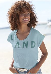 Helles blaues T-Shirt mit kurzen Ärmeln und fettem grünen Aufdruck mit der Aufschrift "SAND". Weicher Stoff und lässiger Schnitt, kombiniert mit einem gewebten Gürtel und Jeansshorts.