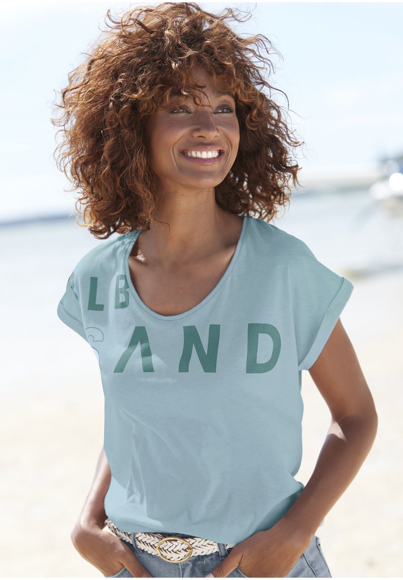 Helles blaues T-Shirt mit kurzen Ärmeln und fettem grünen Aufdruck mit der Aufschrift "SAND". Weicher Stoff und lässiger Schnitt, kombiniert mit einem gewebten Gürtel und Jeansshorts.