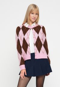 Rožinė ir ruda argyle rašto kardiganas su apvalia apykakle, sagų uždarymu ir raukšlėtomis rankovėmis, dėvimas ant baltos marškinių ir tamsiai mėlynos sijono.