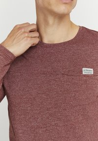 Mørkerød langermet skjorte laget av teksturert stoff, med en rund hals og et lite, rektangulært logo-merke på brystet.