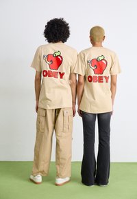 Beige bavlněné trička s grafickým motivem červeného jablka a textem "OBEY" na zádech. Jeden model má na sobě béžové cargo kalhoty, druhý tmavé džíny.