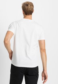 T-shirt bianca a maniche corte realizzata in morbido cotone, con scollo rotondo e orlo dritto, indossata con pantaloni neri, vista da dietro.