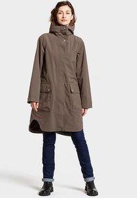 Bruine waterdichte jas met capuchon, voorzien van drukknoopsluitingen en twee voorzakken. Gecombineerd met een blauwe spijkerbroek en zwarte enkellaarsjes.