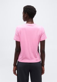 Άτομο με κοντά σγουρά μαλλιά, φοράει ροζ κοντομά νικο πουκάμισο και μαύρο παντελόνι, στέκεται με την πλάτη προς τα έξω μπροστά σε ένα απλό φωτεινό φόντο.