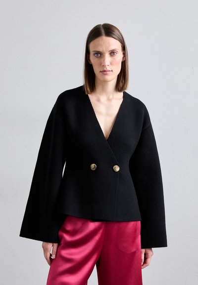 Chaqueta negra tipo envoltura con un escote en V profundo, mangas oversized, doble botón dorado, combinada con pantalones de satén rosas. Textura suave, diseño moderno.