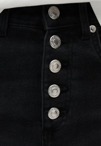 Κοντινό πλάνο μαύρων τζιν από denim με πέντε ασημένια κουμπιά χαραγμένα με τις λέξεις "ONLY BLUE DENIM" και "1995" κατά μήκος της μπροστινής πλάκας.