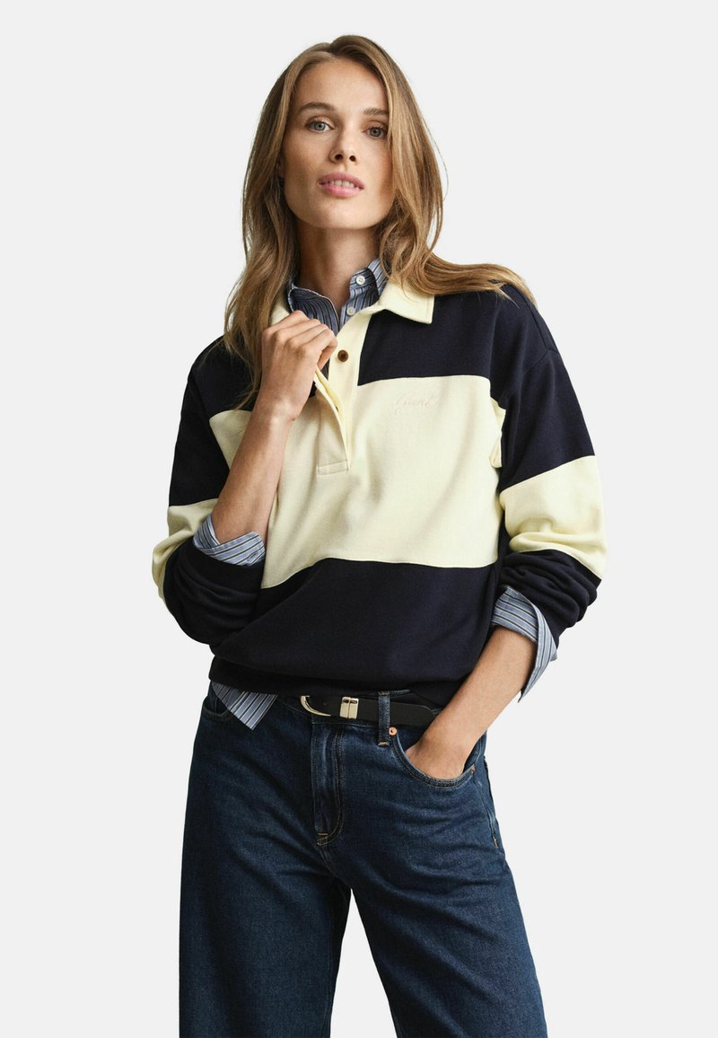 Marineblauwe en crèmekleurige color-blocked pullover met lange mouwen en een kraag. Draag je over een lichtblauw gestreept shirt. Gecombineerd met donkere spijkerbroeken.