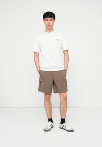 Νεαρός άνδρας στέκεται με τα χέρια στις τσέπες, φορώντας λευκό μπλουζάκι, καφέ cargo σορτς, μαύρες κάλτσες και λευκά αθλητικά παπούτσια πάνω σε απλό φόντο.