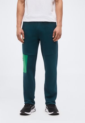 Mann trägt dunkel petrolfarbene Hose mit grünem Seitentaschen und schwarzen Sneakers, steht vor einem einfarbigen hellen Hintergrund.