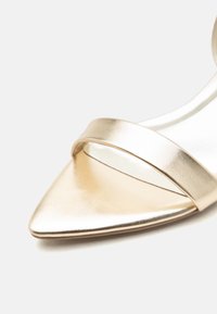 Χρυσό μεταλλικό πέδιλο με μυτερή μύτη και ψηλό τακούνι, με ένα φαρδύ λουρί μπροστά, σε λευκό φόντο.