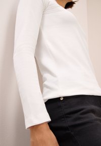 Haut blanc à manches longues avec une texture lisse et une coupe ajustée, associé à un pantalon noir avec un discret détail de bouton métallique.