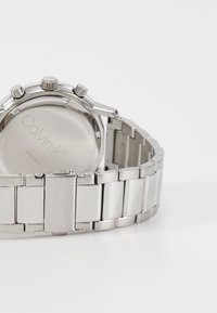 Skrivbordsur i silverfärgad rostfritt stål med metallarmband, chronograph-funktioner och polerad yta. Baksidan märkt med "Calvin Klein."