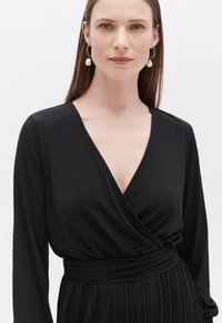 Schwarzes Wickelkleid mit langen Ärmeln, V-Ausschnitt und plissiertem Rock. Glattes Material und definierter Taillenbund. Perlen-Ohrringe als Akzent.