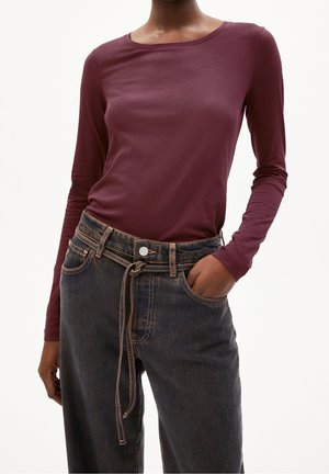 Personne portant une chemise bordeaux ajustée à manches longues, rentrée dans un jean noir délavé taille haute avec une ceinture en corde, une main dans la poche.