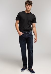 Młody mężczyzna stojący, ubrany w czarną koszulkę, ciemnoniebieskie dżinsy i granatowe trampki z brązowymi sznurowadłami, na jednolitym, jasnym tle.