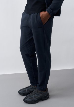 Mees, kes kannab tumedate triibuliste küljetaskutega pükse, musti tossusid ja musta jopet, seisab ühe käega taskus.