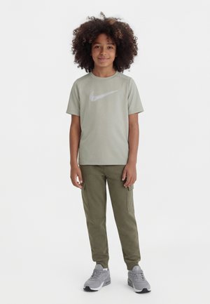 Niño con cabello rizado que lleva una camiseta Nike gris claro, pantalones cargo verde oliva y zapatillas grises, de pie frente a un fondo blanco.