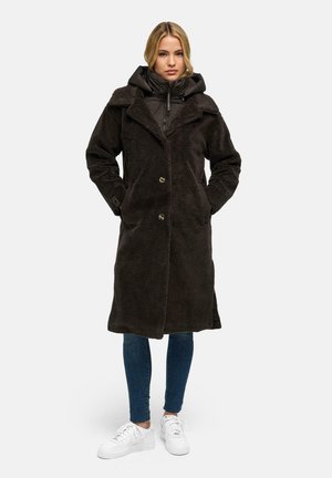 Kvinde iført en lang sort fuzzy frakke over en sort dunjakke, blå jeans og hvide sneakers, stående med hænderne i frakkens lommer.